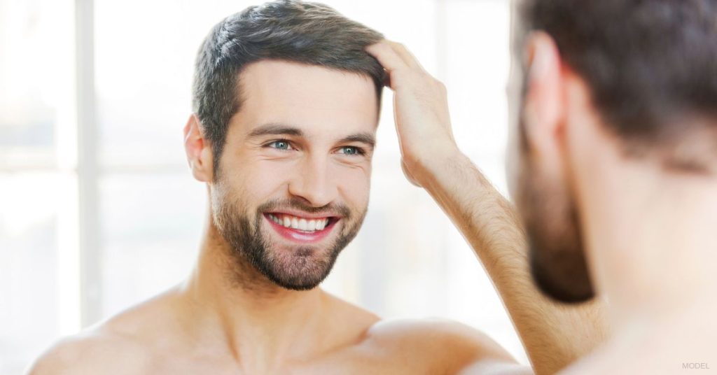 A man smiling at himself in front of the mirror and styling his hair. (Model)